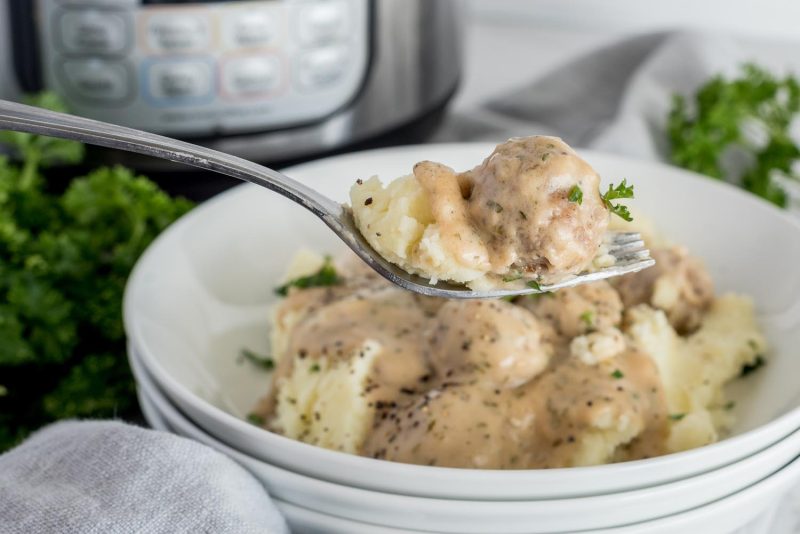 A fork with two Swedish Meatballs in the foreground and a bowl of mashed potatoes topped with Swedish Meatballs in the background.