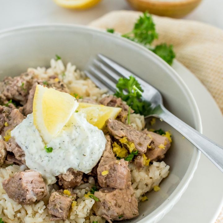 A bowl served with Instant Pot Greek pork over lemon rice, topped with tzatziki sauce and lemon wedges, with a bowl of tzatziki sauce in the background.