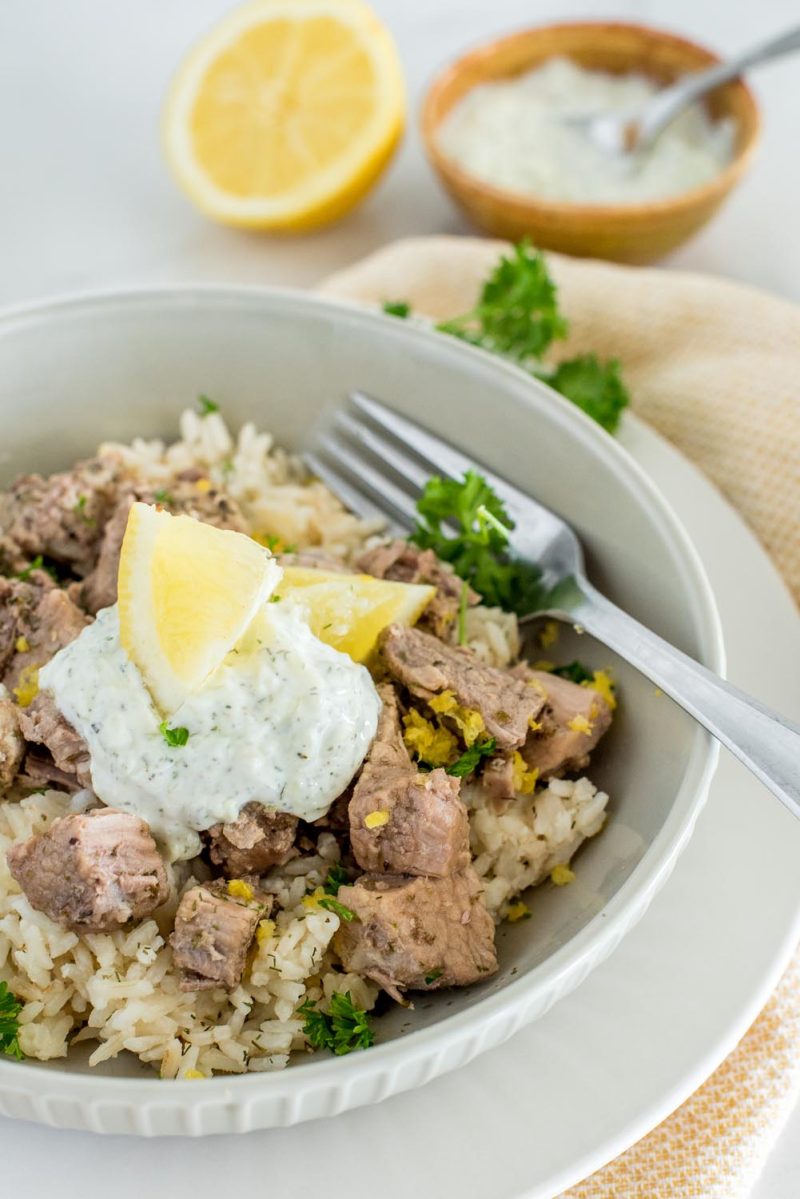 A bowl served with Instant Pot Greek pork over lemon rice, topped with tzatziki sauce and lemon wedges, with a bowl of tzatziki sauce in the background.