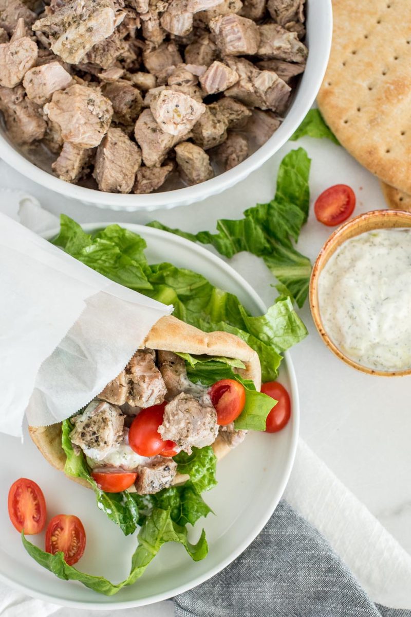 Overhead picture of Instant Pot Greek pork made into a taco with lettuce, tomato, and tzatziki sauce in a pita, with a serving bowl of Greek Pork and a bowl of tzatziki sauce in the background.