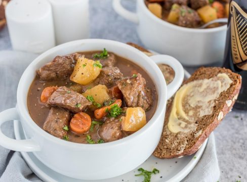 Instant Pot Guinness stew in a white bowl with buttered bread on the side and a bottle of Guinness beer in the background.