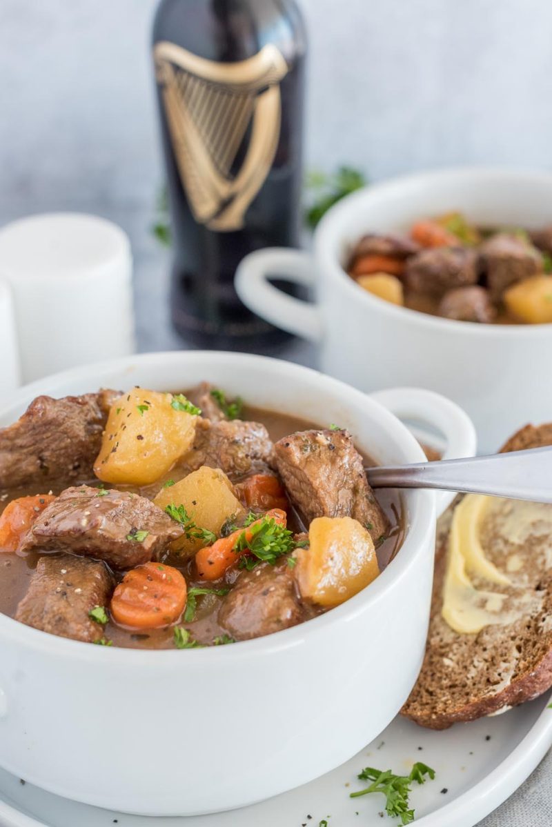 Guinness Stew in a white bowl with buttered bread on the side and a bottle of Guinness beer in the background.