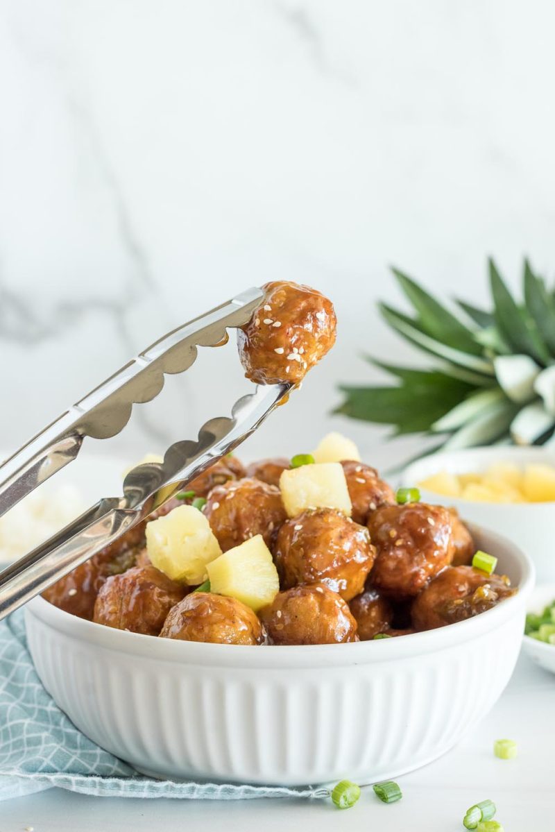 A serving bowl with Hawaiian meatballs that were cooked in an Instant Pot and tongs holding a single meatball in the air.