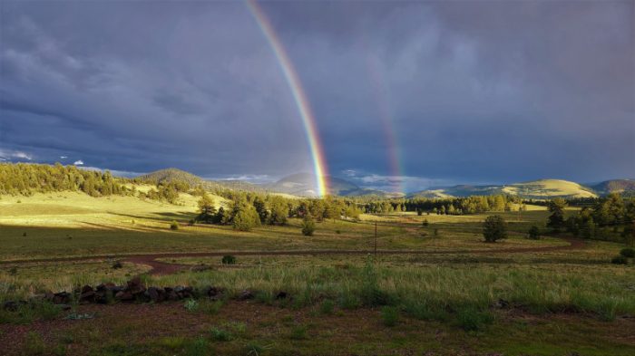 A wide landscape shot, with land in the bottom half and a cloudy sky in the top half, with a double rainbow appearing to come down right in the center of the landscape. Photo by Laurie Bratten.