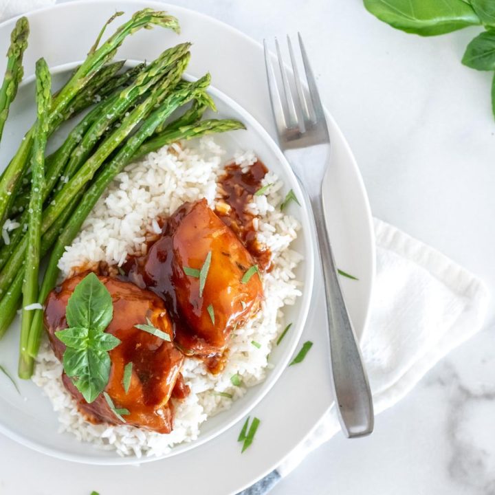 An overhead vertical photo of a white plate and silver fork on a white background. The white plate is in the center left side, filled with cooked asparagus, a bed of white rice, and topped with honey garlic chicken.