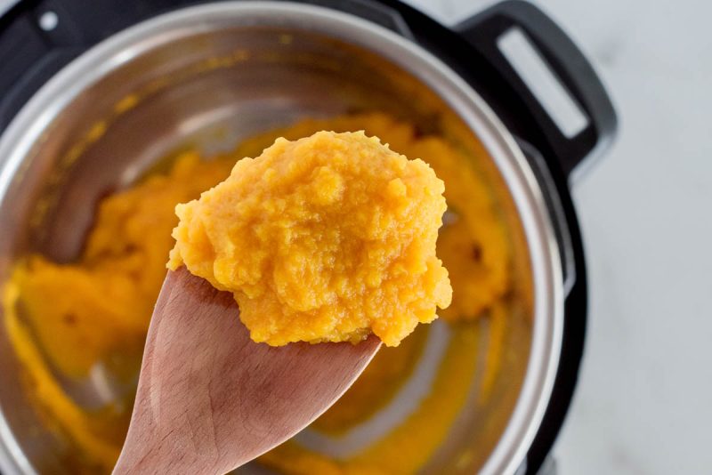Pumpkin puree on a wooden spoon over an Instant Pot with freshly prepared pumpkin puree.