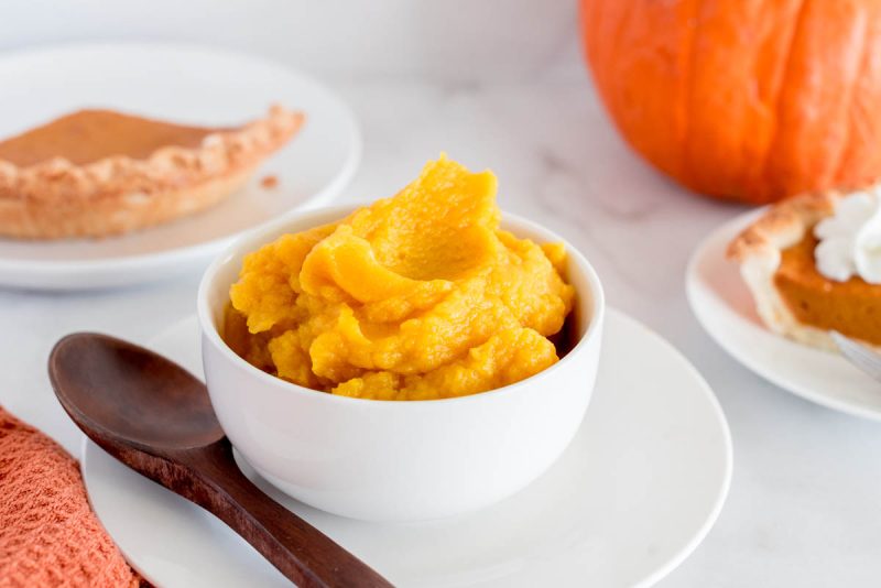 Instant Pot pumpkin in a white bowl with a pumpkin pie off to the side and a pumpkin in the background.