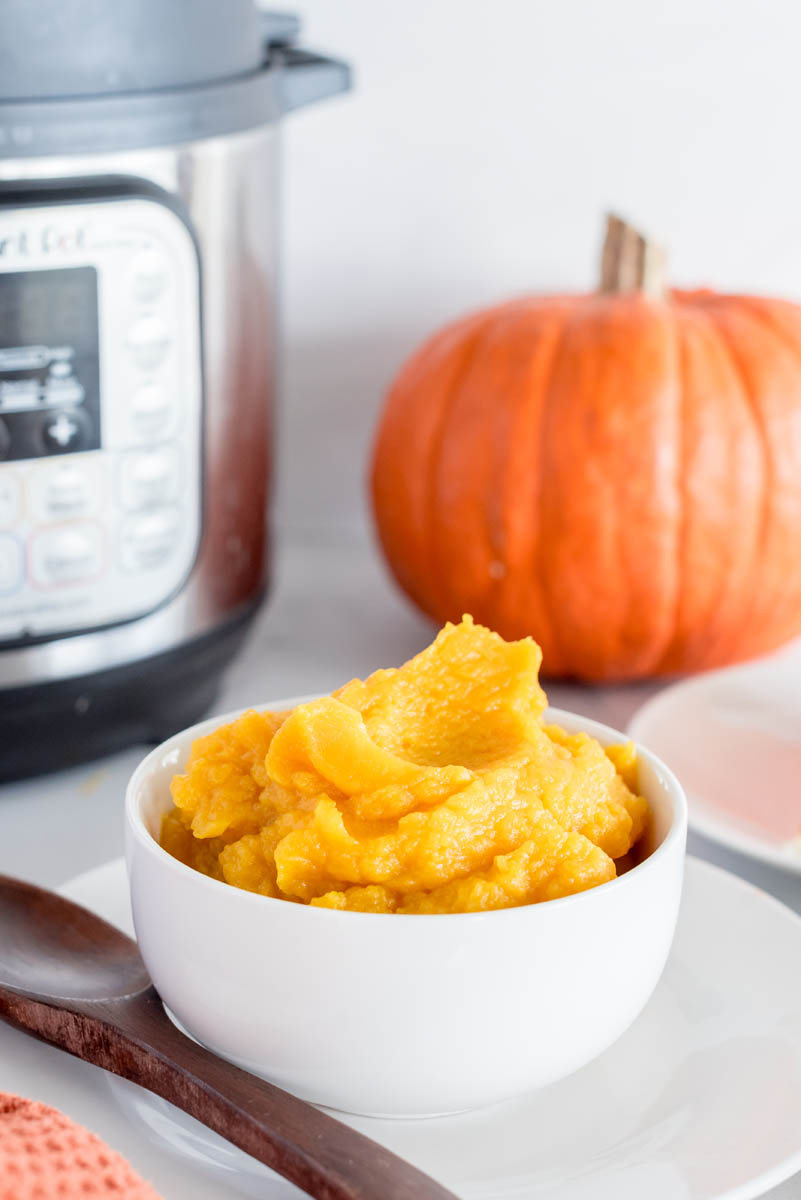 Pumpkin puree in a white bowl with a pumpkin and an Instant Pot in the background.