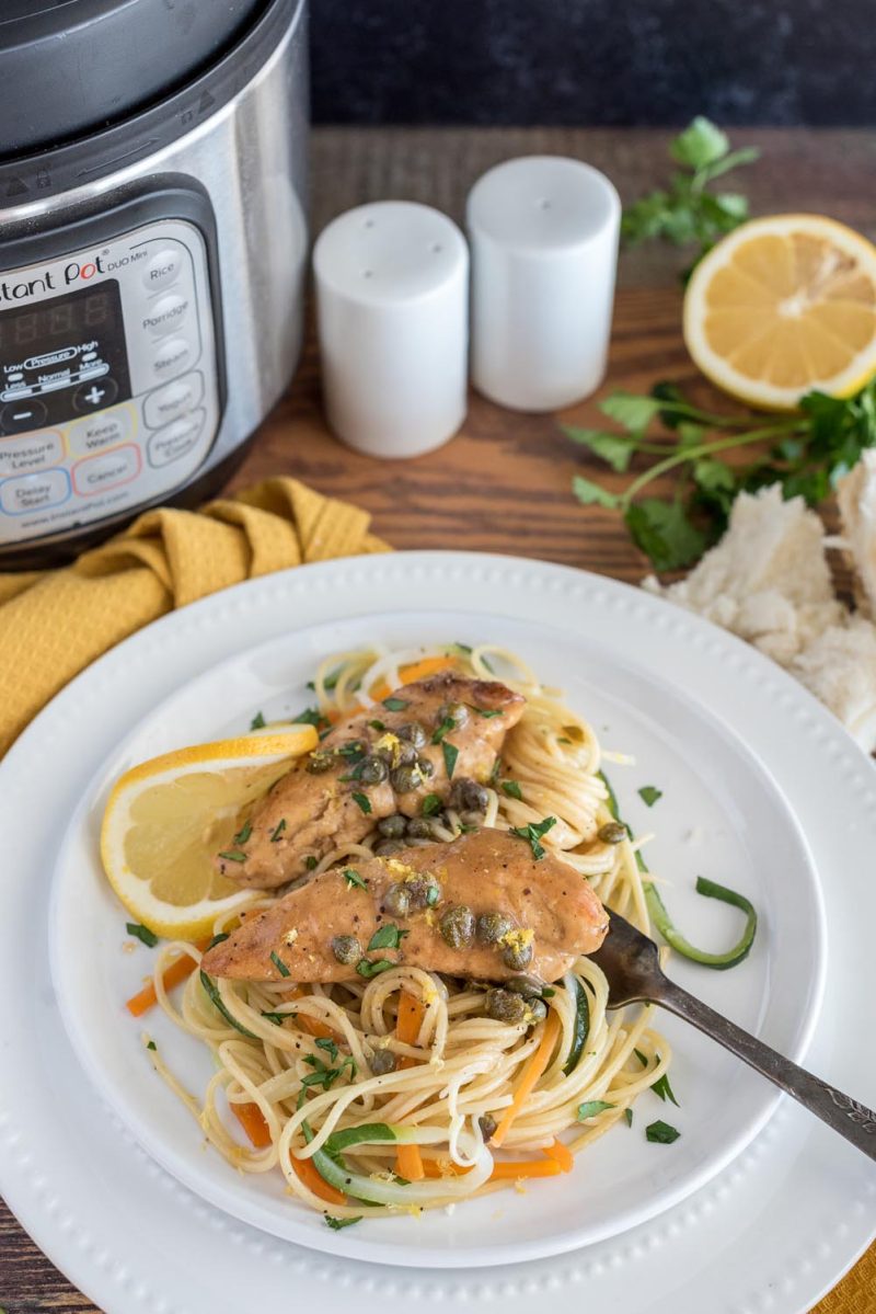 Picture of lemon chicken piccata served over pasta, carrots, and zucchini, placed on a white plate in front of an Instant Pot.