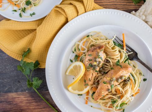 Instant Pot lemon chicken piccata on a white plate with a fork, served over pasta, carrots, and zucchini, with a lemon slice on the side.