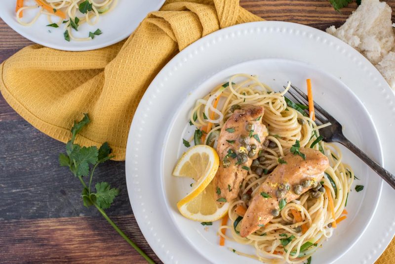 Instant Pot lemon chicken piccata on a white plate with a fork, served over pasta, carrots, and zucchini, with a lemon slice on the side.