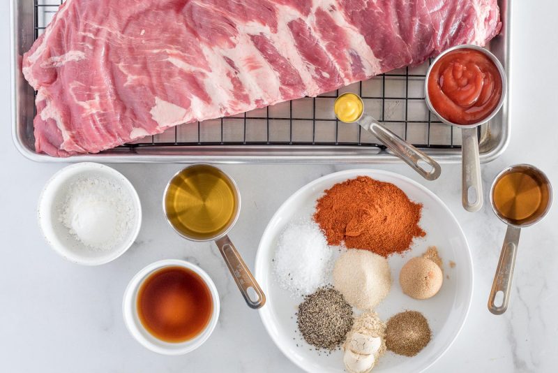 Ingredients including St. Louis ribs on a black metal rack, and metal measuring spoons of ketchup, Worcestershire sauce, spices, cornstarch, and mustard.