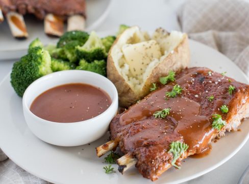 A picture of Instant Pot Memphis style ribs plated with extra homemade BBQ sauce, a baked potato, and steamed broccoli, with more plated ribs in the background.