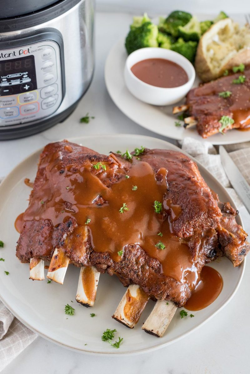 A picture of a white plate with about half a rack of Memphis style ribs smothered in homemade BBQ sauce, a plate with ribs, baked potato and broccoli in the background, all placed next to an Instant Pot.