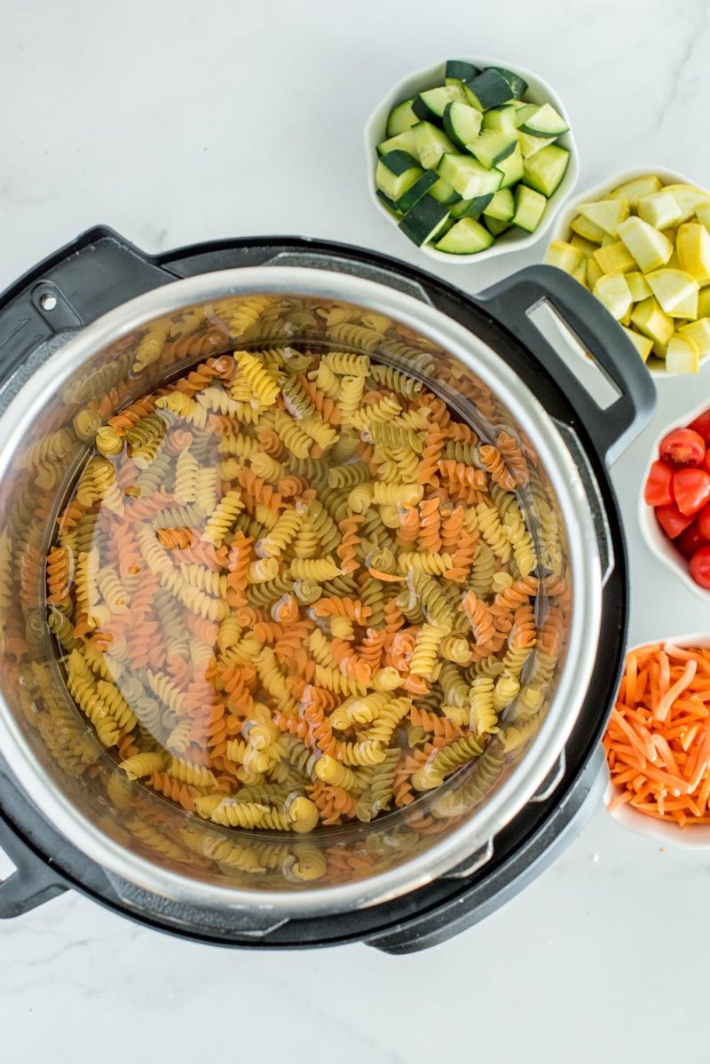 Rotini noodles in an Instant Pot, with bowls of cucumber, yellow squash, cherry tomatoes, and matchstick carrots for making pasta salad.
