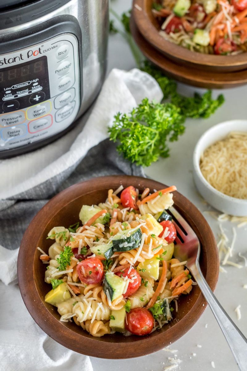 Picture of Instant Pot pasta salad with rotini, sliced cherry tomatoes, zucchini, cucumber, parmesan, and fresh parsley, in a wooden bowl, placed in front of an Instant Pot.