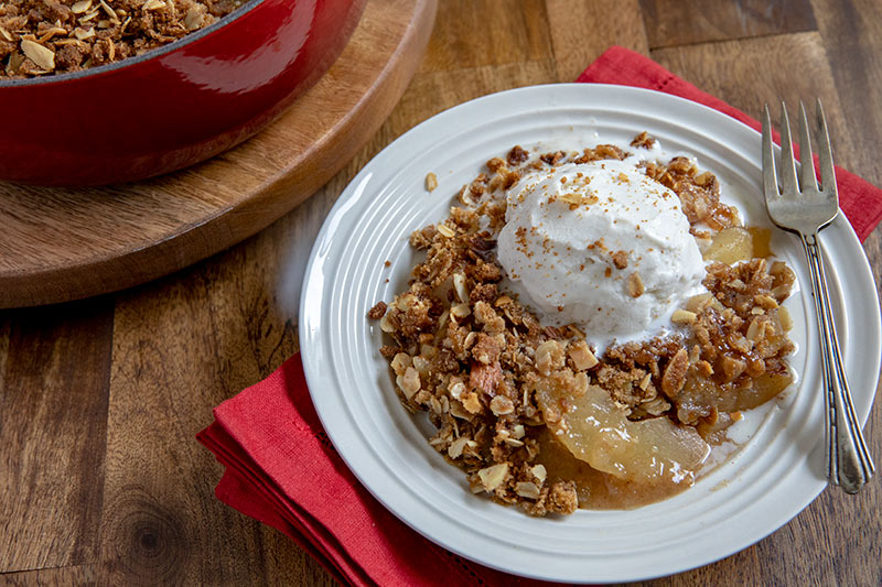 45 degree shot of apple crisp on a white plate, with a big dollop of whipped cream and a toasted topping visible on a white plate
