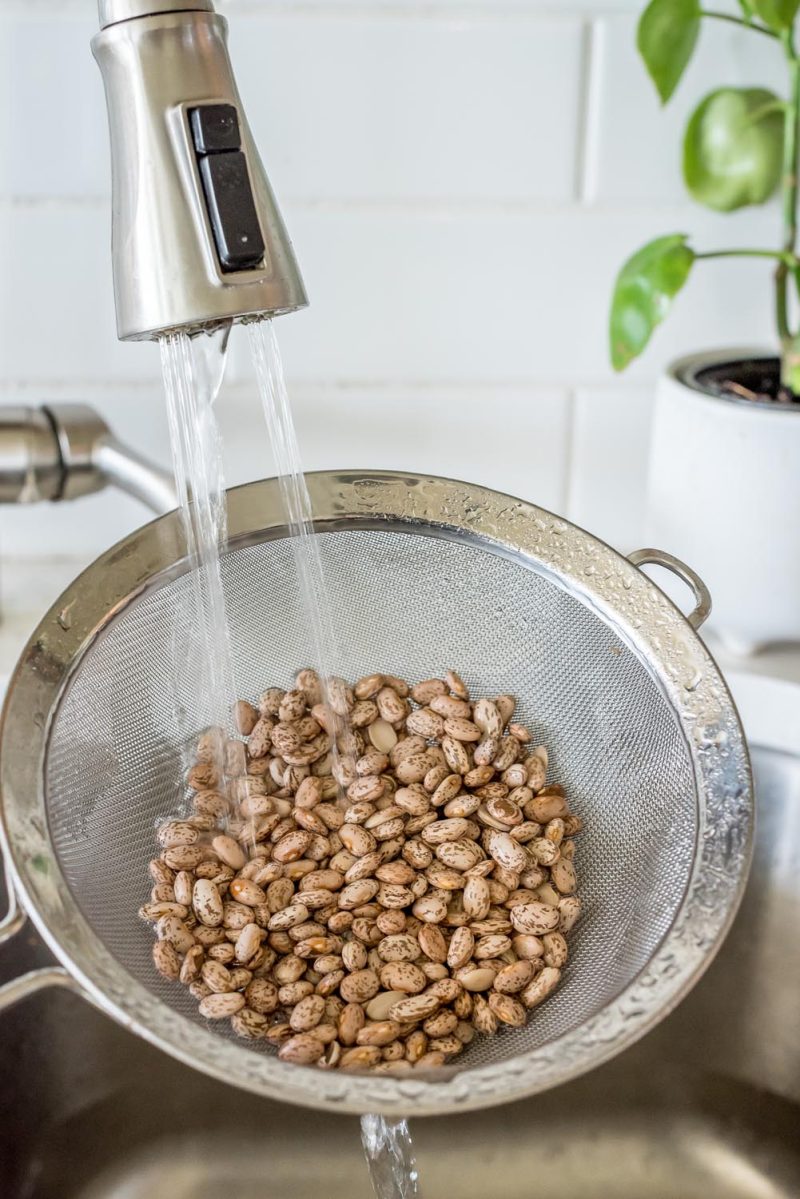 Rinsing dried pinto beans in a wire mesh strainer in a sink.