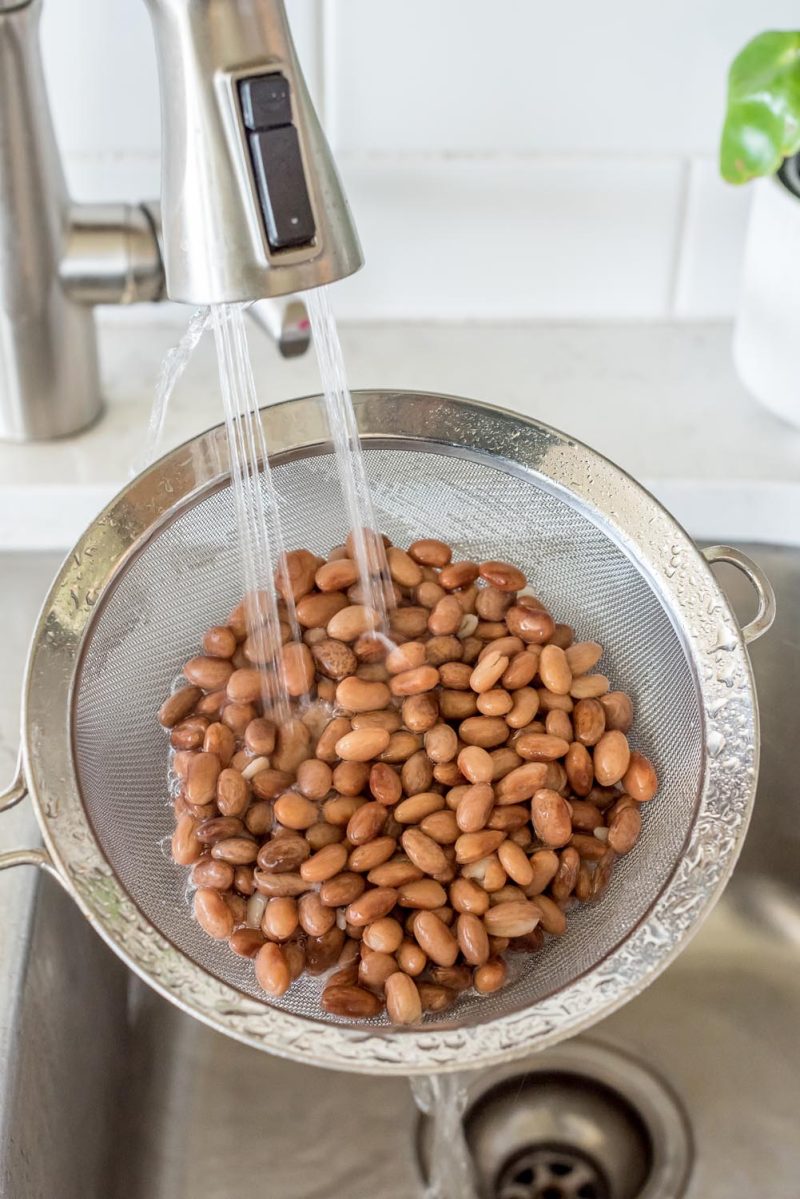 Rinsing soaked pinto beans in a wire mesh strainer in a sink.