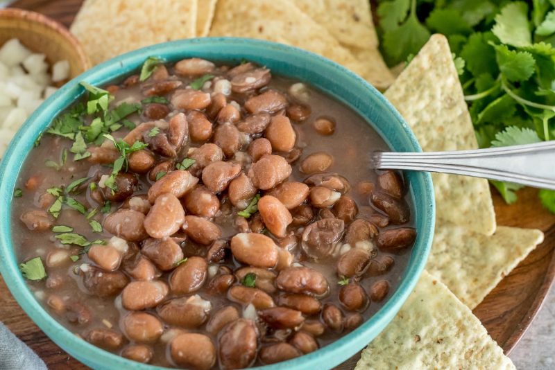 A green bowl with Instant Pot pinto beans, topped with diced onion and cilantro, with chip, a bowl of diced onions, and cilantro in the background.