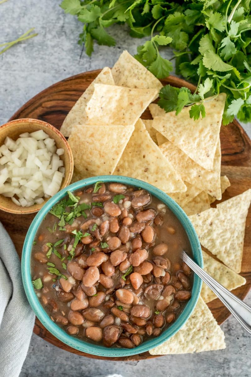 An overhead picture of a green bowl with Instant Pot pinto beans, topped with diced onion and cilantro, with chips and a bowl of diced onions in the background.