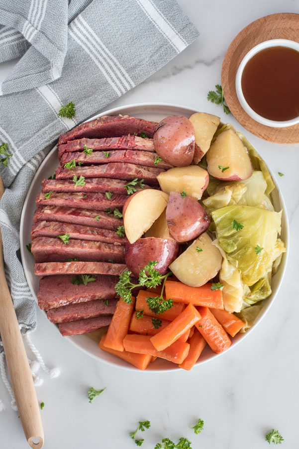 Overhead of a white serving platter filled with Instant Pot Corned Beef, cabbage, potatoes, and carrots with flavorful sauce on the side on a white background with a grey an white linen napkin.