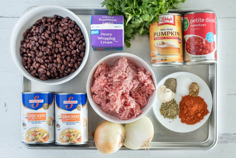 an overhead shot of the ingredients needed to make pork and black bean soup, including ground pork, chicken broth, black beans, heavy cream, onion, cilantro, pumpkin, tomatoes, and spices