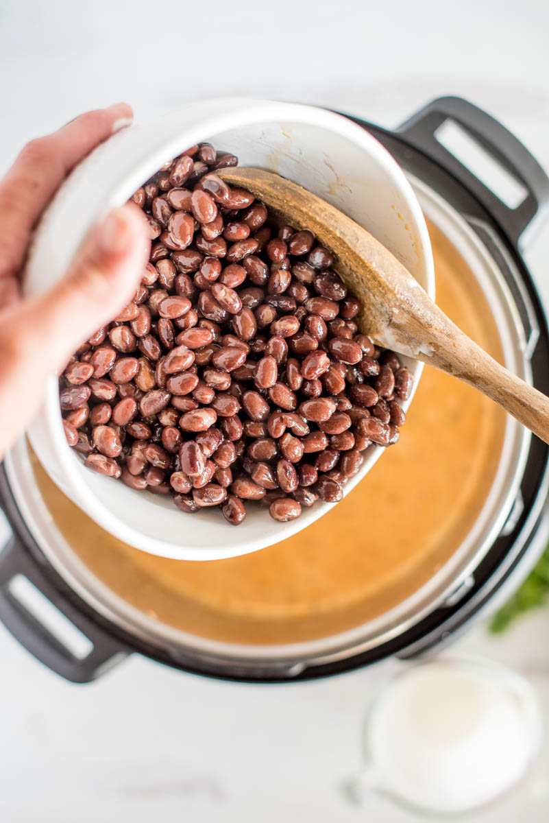 an overhead shot of a hand holding a white bowl of black beans and using a wooden spoon to scrape them in the Instant Pot below the bowl.