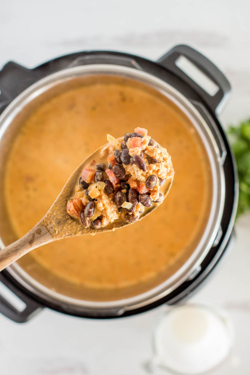 A wooden spoon filled with a heaping scoop of black beans, ground pork, and tomatoes, suspended over an Instant Pot filled with a creamy orange broth.