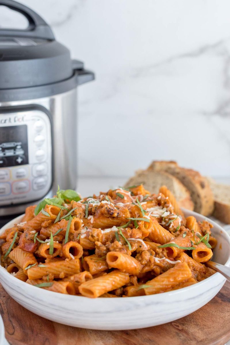Rigatoni pasta with sausage in a white serving bowl and topped with cheese and fresh basil, placed in front of an Instant Pot.