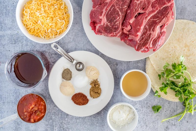 an overhead horizontal shot of the ingredients needed to make beef enchiladas. From top to bottom: shredded Mexican cheese in a white dish, a large plate filled with a well marbled cut of chuck roast, a glass measuring cup filled with beef broth, a small white plate with six piles of seasonings and a measuring spoon, a small white bowl filled with oil, flour tortillas garnished with fresh cilantro, a silver measuring cup filled with red salsa, and another small white bowl filled with cornstarch.