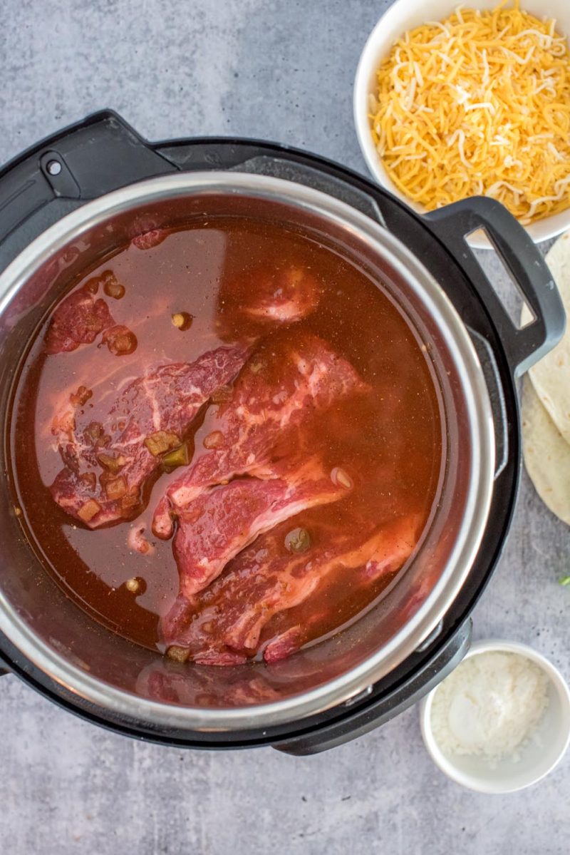 An overhead shot looking into an Instant Pot filled with the red enchilada sauce, salsa and broth, with the marbled chuck roast slightly visible under the liquids.
