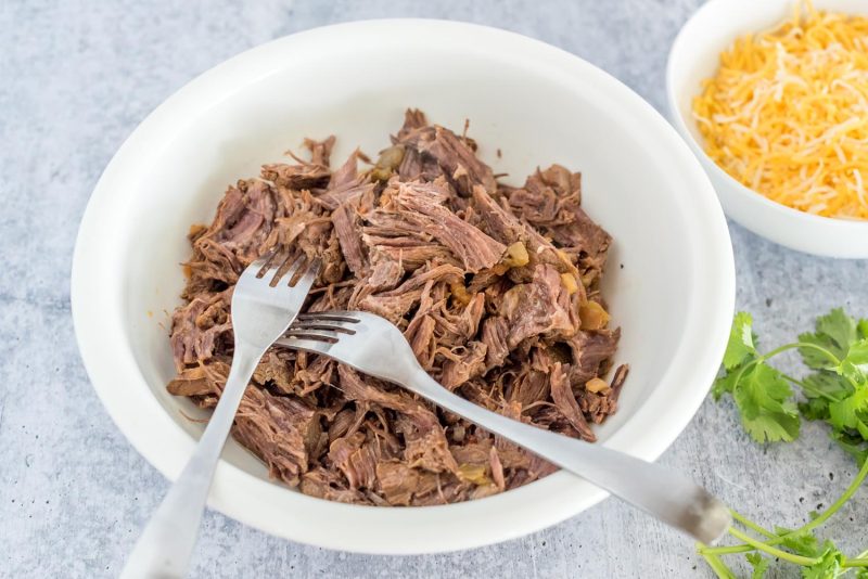 A horizontal photo showing a white bowl with two silver forks used to shred the beef chuck roast, with a small bowl of shredded cheddar cheese in the background.