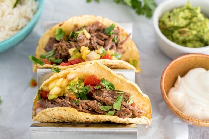 A 45 degree above photo of a silver metal taco holder, holding two small corn tortilla, filled with shredded beef, diced tomatoes, and garnished with cilantro.