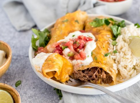 A close-up, horizontal photo of two beef enchiladas, one with a bite taken out so you can see the shredded beef, garnished with sour cream and tomato salsa with shredded lettuce, rice, and lime.