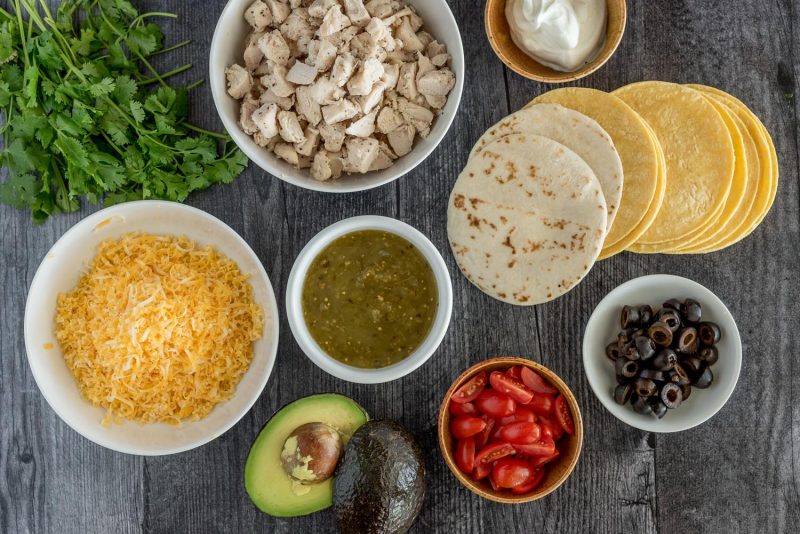 Ingredients for Instant Pot staked enchiladas, including, shredded cheese, enchilada sauce, tomatoes, avocado, olives, tortillas, chicken, and sour cream.