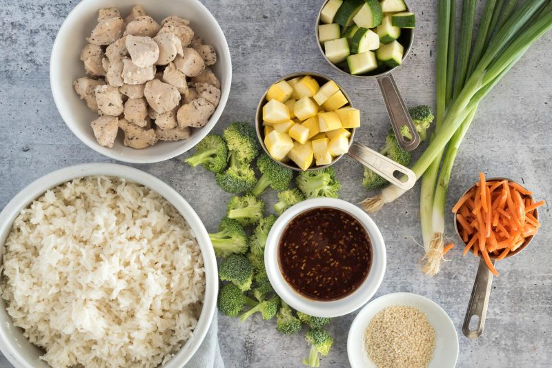 Ingredients for Instant Pot chicken teriyaki bowls including cooked rice, cooked chicken, teriyaki sauce, sesame seeds, carrots, green onion, broccoli, yellow squash, and zucchini.