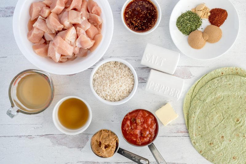 Ingredients shot for Instant Pot Thai chicken wraps, including spices, chili sauce, broth, sesame oil, rice, chicken salsa, peanut butter, butter, and wraps.