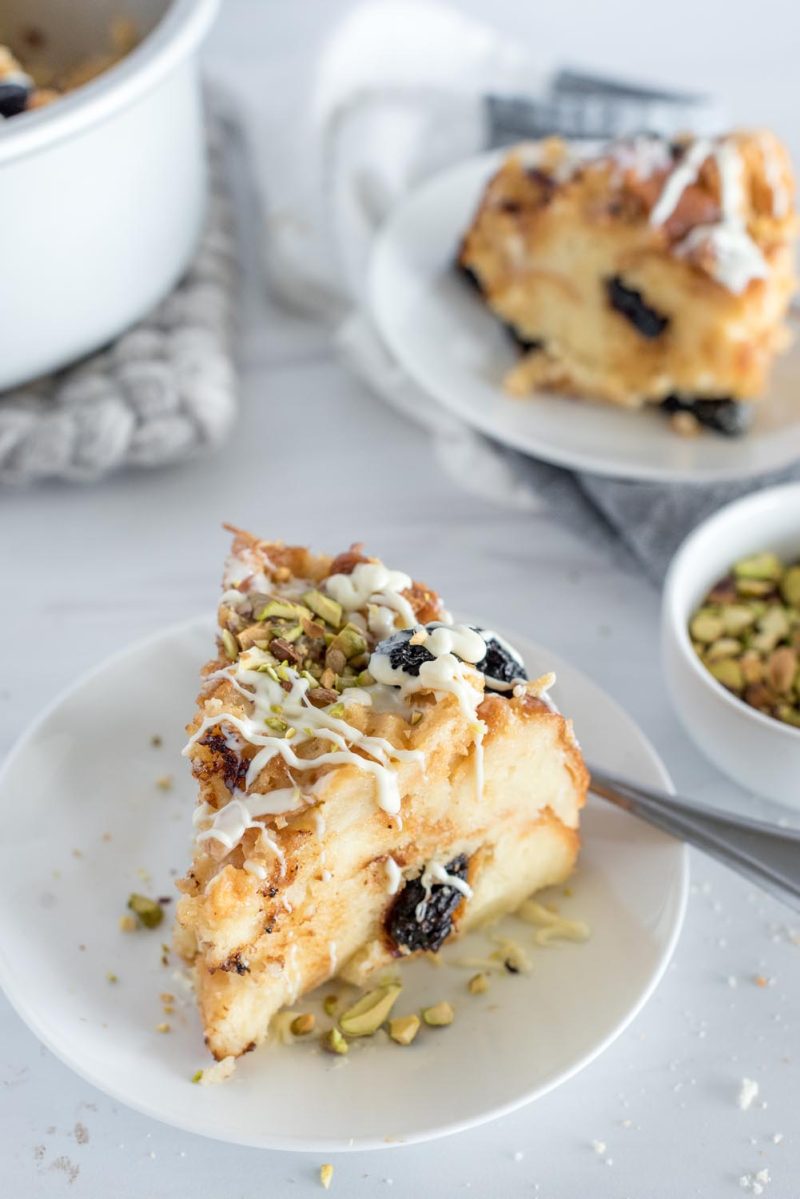 Overnight Instant Pot bread pudding with white chocolate and cherries on a white plate, iced and topped with chopped pistachios.