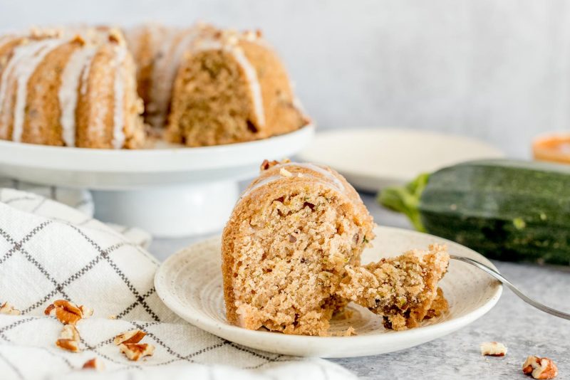 A slice of Insta pot zucchini bread on a white plate with a fork, with a zucchini and the rest of the mini bundt zucchini bread in the background.